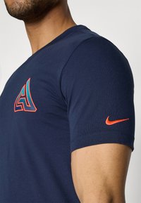 Camiseta de algodón azul marino con un logo geométrico en naranja y turquesa en el pecho y un swoosh de Nike naranja en la manga.