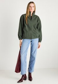 Groene fleece jack met hoge kraag, elastische manchetten en ritssluiting, gedragen met lichtblauwe jeans en bordeauxrode accessoires.