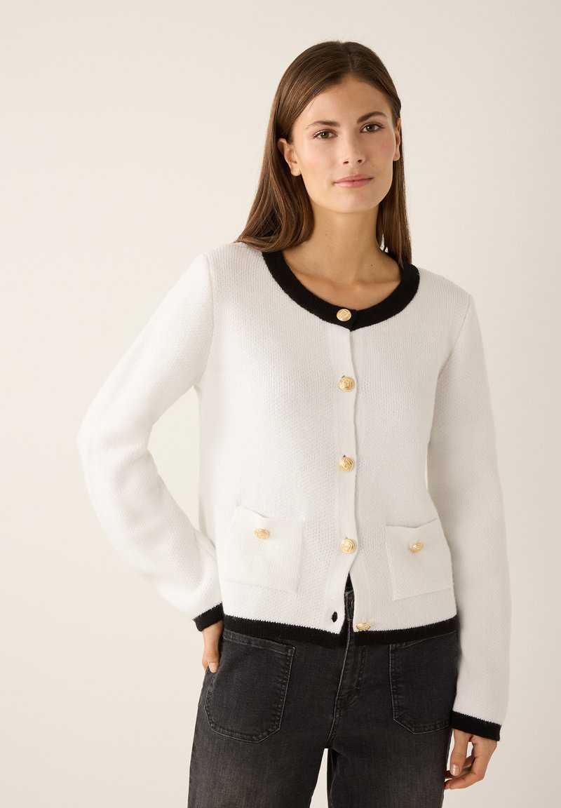 Witte cardigan met zwarte bies, gouden knopen en twee voorzakken. Gebreide textuur met een ronde hals en lange mouwen.