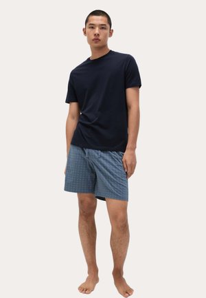 Junger Mann, der barfuß steht und ein einfaches, dunkelblaues T-Shirt sowie kurze, gemusterte blaue Shorts trägt, vor einem schlichten Hintergrund.