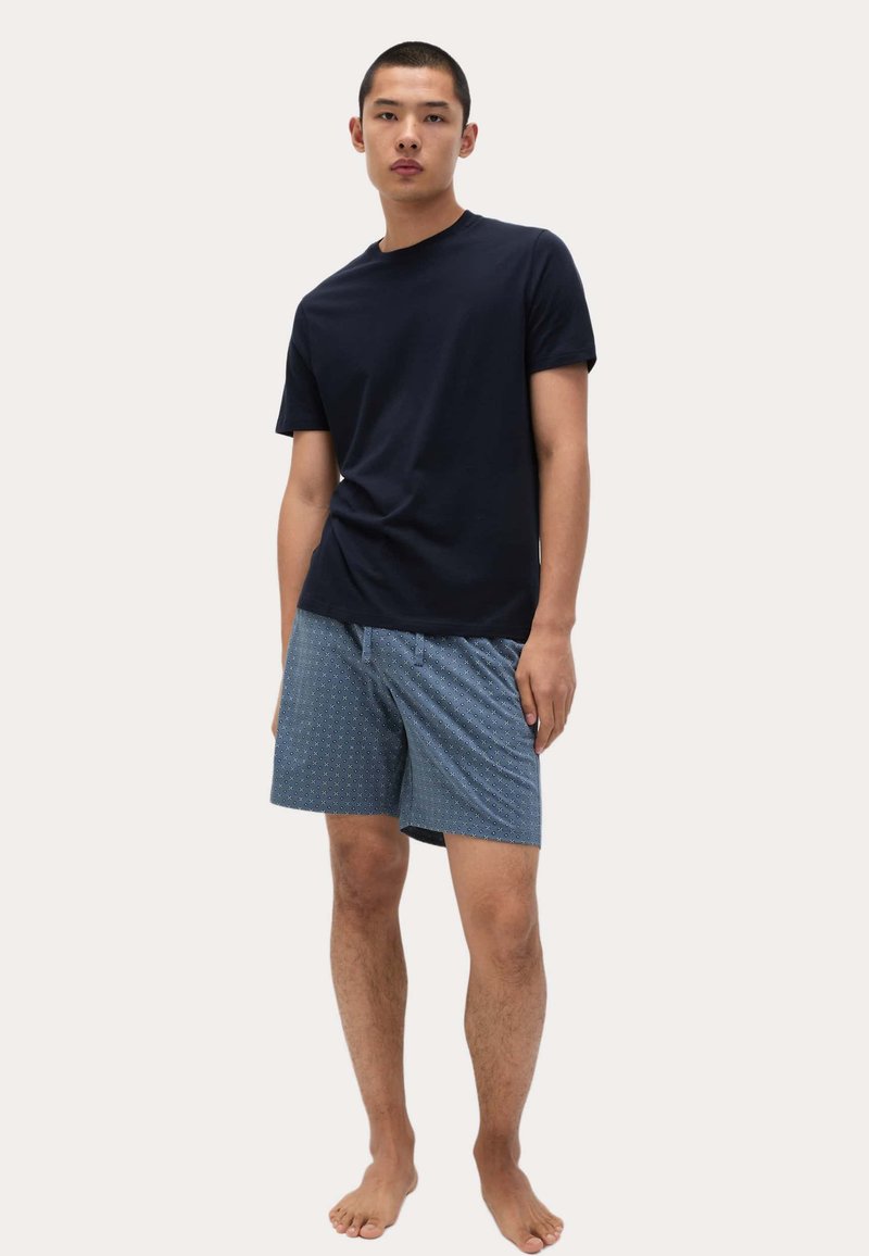 Ung man som står barfota iförd en enkel mörknavy t-shirt och blårandiga shorts framför en enfärgad bakgrund.