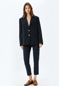 Marineblauwe oversized blazer met een diepe V-hals, twee voorzakken en zilveren knopen, gecombineerd met op maat gemaakte marineblauwe broek en bruine loafers.
