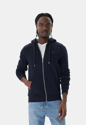 Młody mężczyzna z warkoczami, ubrany w granatową bluzę rozpinaną na zamek na białą koszulę oraz jasnoniebieskie dżinsy, stojący z jedną ręką w kieszeni.