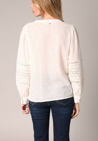 Blouse beige clair à manches longues, avec des détails en dentelle brodée sur les manches, une coupe décontractée et un ourlet arrondi.