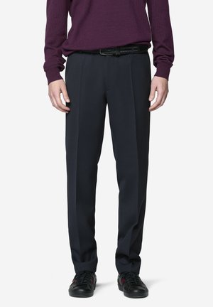 Mann trägt schwarze maßgeschneiderte Hose, schwarzen Gürtel, schwarze Sneaker mit roten Streifen und ein violettes Langarmshirt und steht.