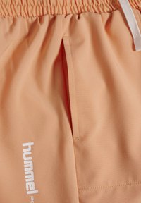Ferskenfarvede shorts med elastisk talje, sidelomme, hvid snor og hvidt "hummel"-logo trykt lodret nær lommen.