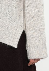 Light gray knitted sweater with ribbed cuffs and side slits, featuring a soft texture and slightly loose fit, worn over a dark garment.