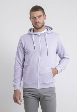 Homme aux cheveux courts et barbe portant un sweat à capuche zippé violet clair et un pantalon gris, debout devant un fond uni.