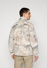Mann som står sidelengs iført en høyhalset fleecejakke med et skogmønster i beige, grått og lyseoransje, samt beige cargo-bukser.