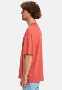 Man met schouderlang bruin haar, gekleed in een losse koraalkleurige T-shirt en lichtblauwe jeans, staand in profiel tegen een effen achtergrond.