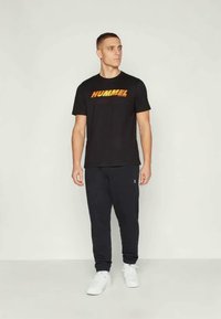 Μαύρη μπλούζα με το πορτοκαλί λογότυπο HUMMEL, συνδυασμένη με ναυτικές παντελόνες και λευκά αθλητικά παπούτσια, σε ουδέτερο φόντο.