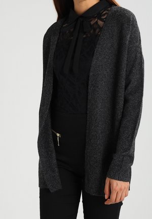 Femme portant un cardigan gris foncé ouvert sur le devant, sur un chemisier en dentelle noire avec un col noué, associé à un pantalon noir avec une fermeture éclair latérale.