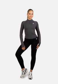 Długi rękaw, sportowa bluzka z zamkiem w ciemnoszarym kolorze, zestawiona z dopasowanymi czarnymi legginsami. Model nosi białe i czarne buty sportowe.