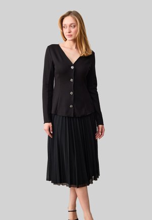 Vrouw met een zwart, lang mouw, blouse met knopen en een V-hals, gecombineerd met een zwarte geplooide midi-rok tegen een grijze achtergrond.