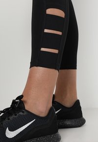 Černé sportovní legíny s výřezy na bocích, které jsou doplněny černými teniskami se stříbrným logem Nike a tečkovanou podrážkou.