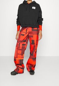 Felpa nera con cappuccio e logo, abbinata a pantaloni rossi con un motivo geometrico e una vestibilità ampia, completata con scarpe nere.