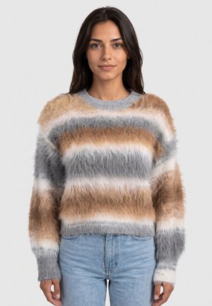 Jonge vrouw met lang, donker haar, gekleed in een gestreepte, zachte trui in beige, grijs en wit en een blauwe spijkerbroek, staand tegen een effen lichte achtergrond.