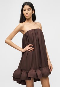 Γυναίκα ντυμένη με αμάνικο, φαρδύ φορέμα σε σκούρο καφέ χρώμα με βολάν στο τελείωμα, ποζάρει με το ένα χέρι στη μέση της μπροστά σε απλό φόντο.