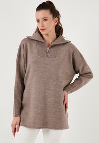 Brauner Strickpullover mit hohem Kragen, halbem Reißverschluss und geripptem Muster am oberen Teil. Besitzt oversized Ärmel und eine lockere Passform.