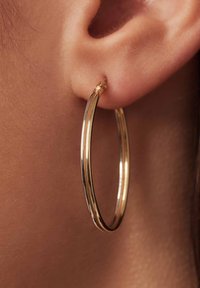Boucle d'oreille en or affichée sur un lobe d'oreille ; présente une surface lisse et brillante avec un design à double couche, mettant en avant une forme ronde.