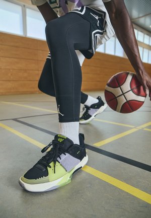 Joueur de basket-dribble avec un ballon rouge et blanc sur un terrain intérieur, portant des leggings noirs et des baskets Jordan multicolores.
