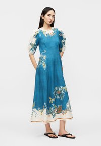 Kobieta w turkusowo-niebieskiej sukience midi z bufkami na rękawach i kwiatowym wzorem, ubrana w czarne klapki, stojąca na jednolitym tle.