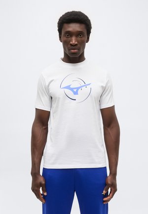 Άνδρας που φοράει λευκό κοντομάνικο μπλουζάκι με μπλε αφηρημένο λογότυπο και μπλε παντελόνι, στέκεται μπροστά σε απλό φόντο.