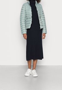 Veste matelassée bleu sarcelle clair, robe côtelée bleu marine et chaussures de sport blanches. La veste présente des détails matelassés et un col montant.