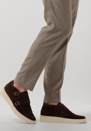 Brown suede shoes with dual buckle accents, round toe, and off-white rubber sole, paired with checkered beige trousers.