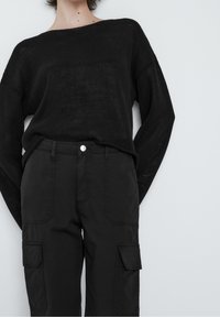 Mustat neulepaidat, joissa on leveä kaula-aukko ja väljä malli, yhdistettyinä mustiin cargo-housuihin, joissa on läpäll�ä varustetut taskut ja nappisulku.