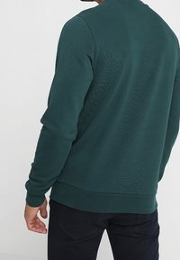 Sudadera de un verde oscuro con cuello redondo, puños y dobladillo acanalados. Textura suave con un acabado liso y detalles de diseño mínimos.