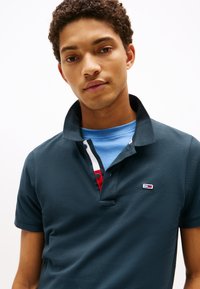 Marineblå polo skjorte i teksturert stoff, med en kontrasterende grå krage og en flerfarget stolpe. Lite logo på venstre side.