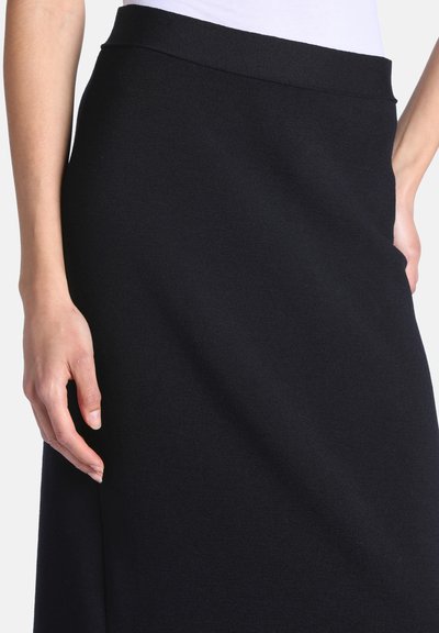 Gros plan d'une personne portant une jupe noire taille haute à texture lisse et un haut blanc rentré.