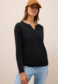 Schwarzes Langarmoberteil mit V-Ausschnitt, das subtile Nähdetails aufweist, kombiniert mit hellblauen Denim-Jeans. Das Material wirkt weich und atmungsaktiv.