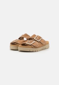 Vaaleanruskeat mokkasiinit, joissa on kaksi säädettävää solkea ja teksturoitu pohja. Kenkien kevyt beige pohja on varustettu kuvioiduilla urilla.