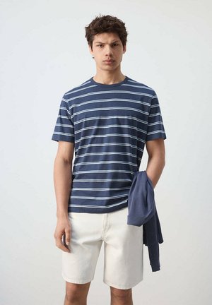 Joven que lleva una camiseta de rayas azules y blancas y pantalones cortos blancos, sosteniendo una sudadera azul marino en su mano izquierda.