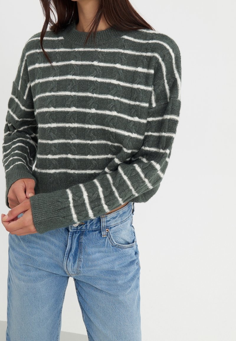 Femme portant un pull en maille torsadée vert avec des rayures blanches horizontales et un jean bleu clair, debout les mains jointes.