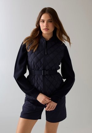 Kobieta z luźnymi falującymi włosami, ubrana w czarną pikowaną bezrękawnik na czarny top z długim rękawem i czarne szorty, stojąca na szarym tle.