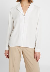 White long-sleeve blouse with a collar, featuring a button-down front and textured, lightweight fabric. Paired with beige trousers.