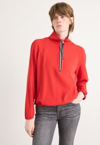 Rode pullover met hoge kraag en halve rits, gemaakt van gladde stof. Heeft elastische mouwen en een subtiele merkdetail. Draagt met grijze jeans.