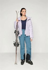 Kobieta w jasnofioletowej kurtce puchowej, niebieskich spodniach narciarskich i czarnych butach narciarskich trzymająca parę nart, stojąca na tle jednolitym.