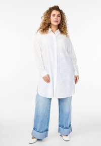 Weiße Bluse mit langen Ärmeln und floralem Muster, Knopfleiste vorne, abgerundeter Saum, kombiniert mit hellen, weiter geschnittenen Jeans und weißen Schuhen.