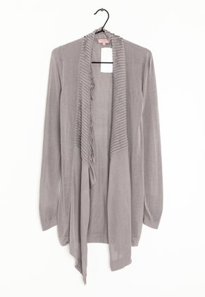 Cardigan grigio con maniche lunghe, texture a coste lungo i bordi frontali, design aperto e un aspetto setoso. Pende da un gancio nero.