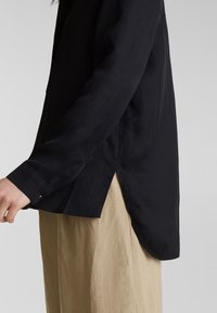 Person in einem langärmeligen schwarzen Shirt mit seitlichem Schlitz und beigen Hosen, von der Taille abwärts vor einem schlichten grauen Hintergrund gezeigt.