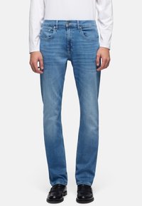 Homme portant un jean droit bleu clair, une chemise blanche à manches longues et des chaussures habillées noires, debout devant un fond uni.