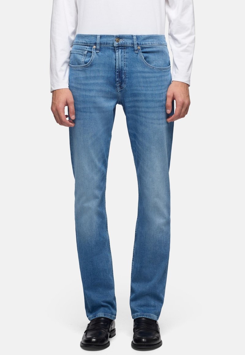 Homme portant un jean droit bleu clair, une chemise blanche à manches longues et des chaussures habillées noires, debout devant un fond uni.