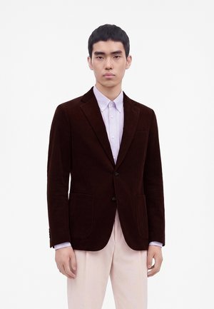 Jeune homme portant un blazer en velours côtelé marron foncé sur une chemise boutonnée violet clair et un pantalon beige clair, debout devant un fond blanc uni.