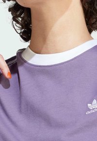 Lila bomulls t-shirt med en vit ribbad halsringning och en liten vit logotyp. Slät textur, korta ärmar, nära vy av halsområdet.