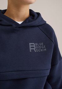 Tumši zils kapuces džemperis ar strukturētu plecu paneli un reljefu "BLUE RIDGE DENIM" logo uz krūtīm. Mīksta auduma, ar brīvu piegriezumu.