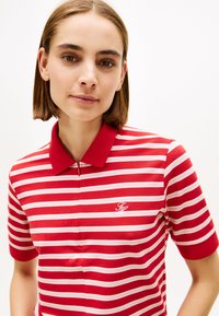 Punaste ja valgete triipudega lühikeste varrukatega polosärk, millel on krae ja tõmblukk. Vasakul küljel on tikitud logo.